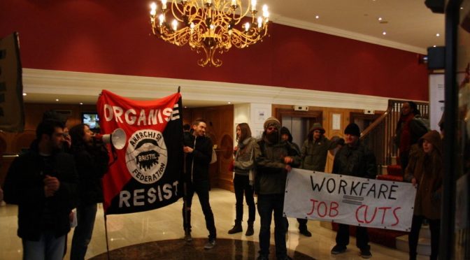 inside a fancy hotel lobby a dozen or so protesters hold up banners saying 'workfare = jobcuts' and 'organise, resist, bristol anarchist federation', one person shouts on a megaphone